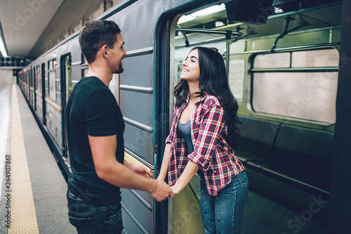 Young man and woman use underground. Couple in subway. Young brunette stand in underground carriage and smile. She hold man's hands. Guy stand on platform. Love stiry. Cheerful.