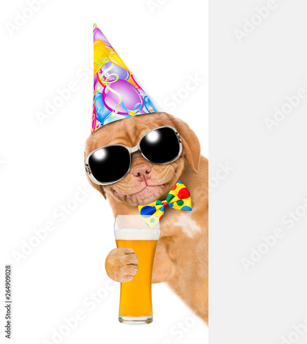 Smiling puppy with sunglasses in birthday hat with light beer behind empty white banner. isolated on white background © Ermolaev Alexandr