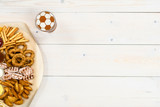 Diverse snacks and beer with a football on a beer foam. Light wooden background. Top view. Empty space for text
