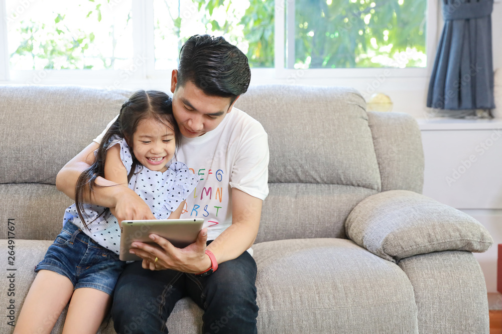 happy asian dad teaching his daughter to use or play tablet on grey sofa in living room with laughing and smiling face (lifestyle with technology concept) happy asian dad teaching his daughter to use or play tablet on grey sofa in living room with laughing and smiling face (lifestyle with technology concept)