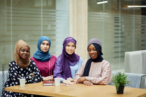 Portrait of smart elegantly dressed caucasian and african muslim female friends look at camera with cheerful expression at office background