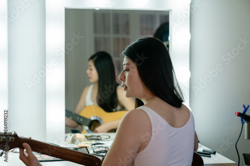 Fototapeta Naklejka Na Ścianę i Meble -  back view beautiful korean woman singer playing acoustic guitar sitting at vanity table in dressing room with applied makeup. young asian girl musician ready for show concert practicing in backstage.