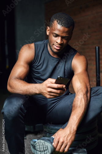 Portrait of african athlete man checking his smart phone while resting after training in gym.