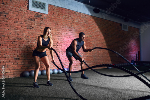 Interracial family couple exercising with battle ropes at gym. Caucasian woman doing battling rope workout training together with cross fit African coach © alfa27