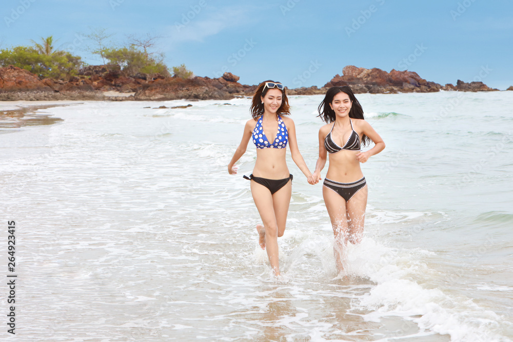 two happy asian sexy women in black and blue bikini who holding hand together and running with smiling and laughing face on beach with clear blue sky on holiday vacation