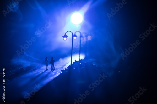 Young couple holding each other hands. Slowly walking under white street lights in night. Dark time. Peaceful atmosphere in mist. Foggy air.