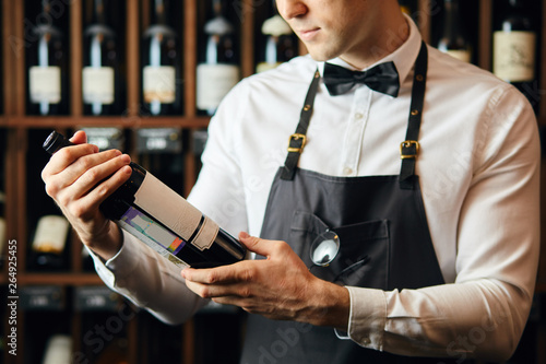 Cropped shot of young caucasian cavist dressed in white shirt and bowtie working in big vine shop presenting a bottle of red wine to customer