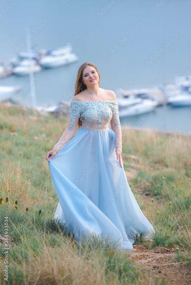 A girl with long hair on the beach. She's wearing a beautiful blue dress . In the background view of the Bay, sea and city. Soft focus. A girl with long hair on the beach. She's wearing a beautiful blue dress . In the background view of the Bay, sea and city. Soft focus.