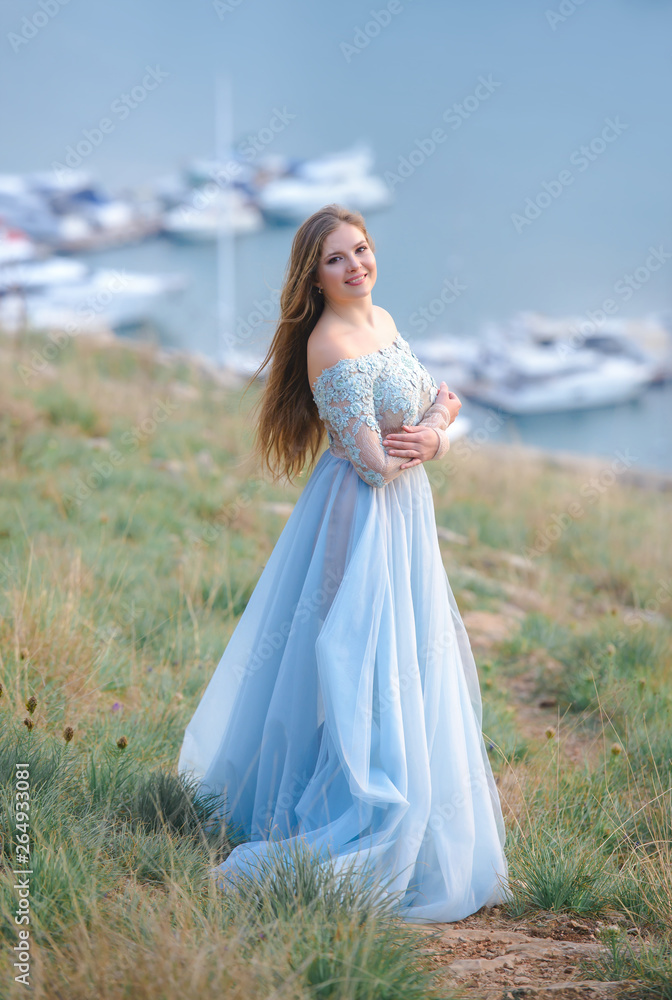 A girl with long hair on the beach. She's wearing a beautiful blue dress .    In the background view of the Bay, sea and city. Soft focus.