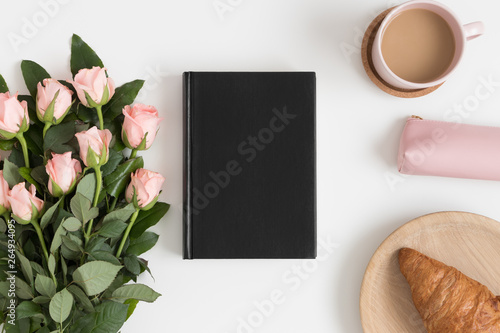 Top view of a black book mockup with a bouquet of pink roses, coffee and a croissant on a white table.