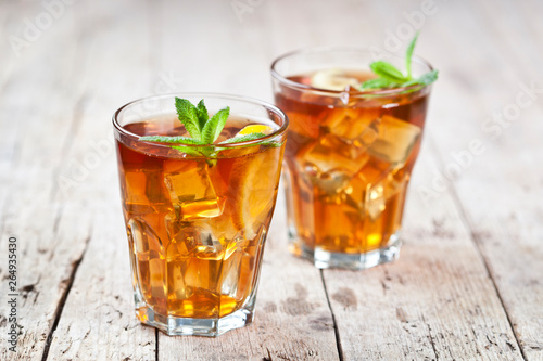 Two glasses with traditional iced tea with lemon, mint leaves and ice cubes in glass on rustic wooden table.