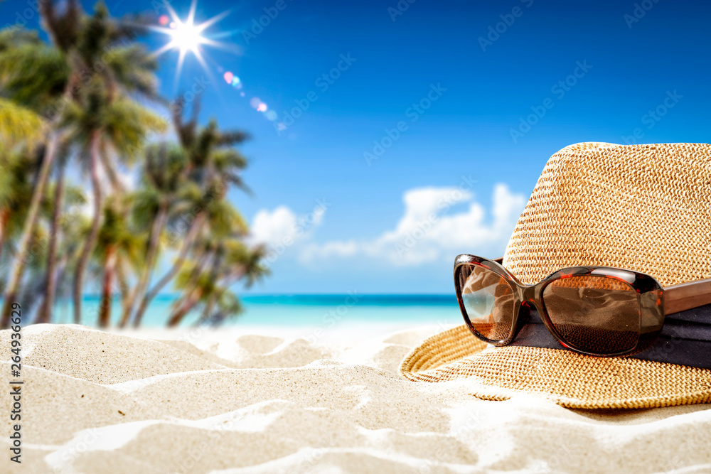 Summer sunglasses with hat on sand and blue towel. Free space for your decoration. Blurred background of beach with palms and ocean Summer sunglasses with hat on sand and blue towel. Free space for your decoration. Blurred background of beach with palms and ocean