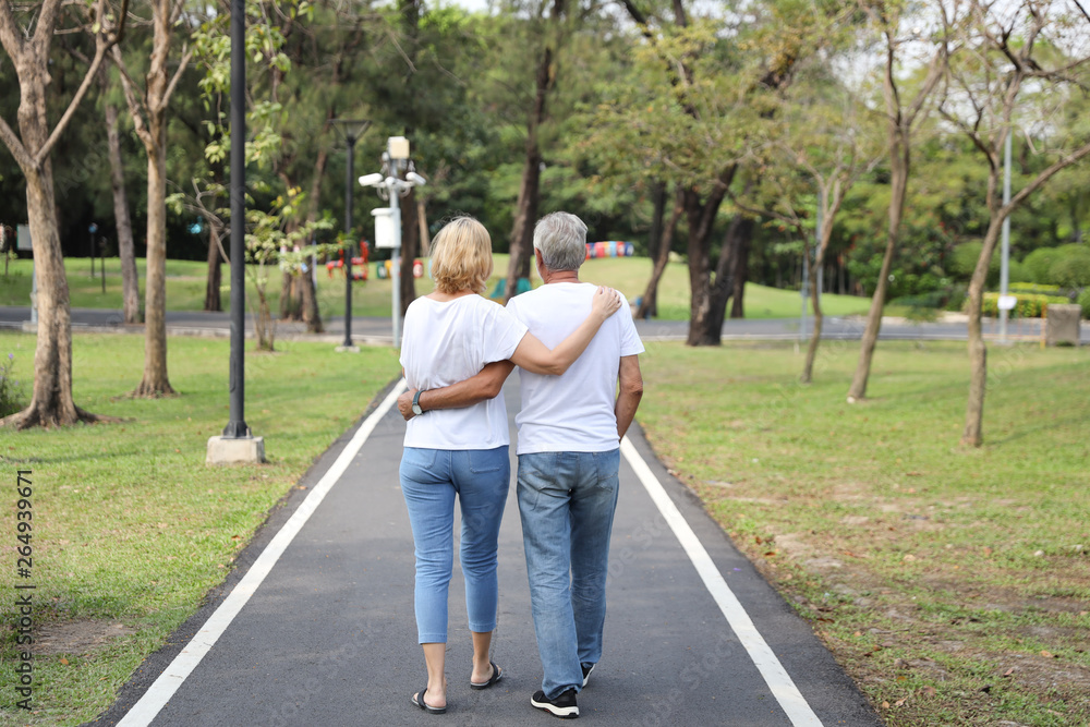 back view elderly couple with white shirt and blue jean embracing and walking in park in summer time back view elderly couple with white shirt and blue jean embracing and walking in park in summer time