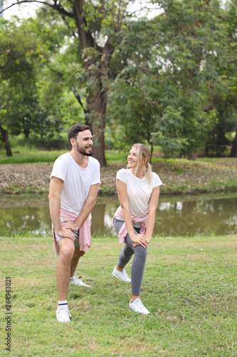 image of young caucasian couple enjoying in exercising and stretching outdoor with smiling faces and green trees during summer time in the park