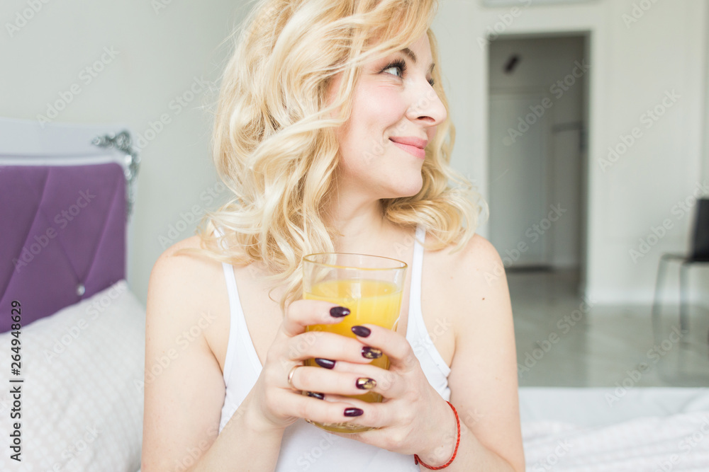 Attractive blonde woman sits on the edge of the bed at home and holds a stocker with orange juice in her hand. Morning mood and vigor Attractive blonde woman sits on the edge of the bed at home and holds a stocker with orange juice in her hand. Morning mood and vigor