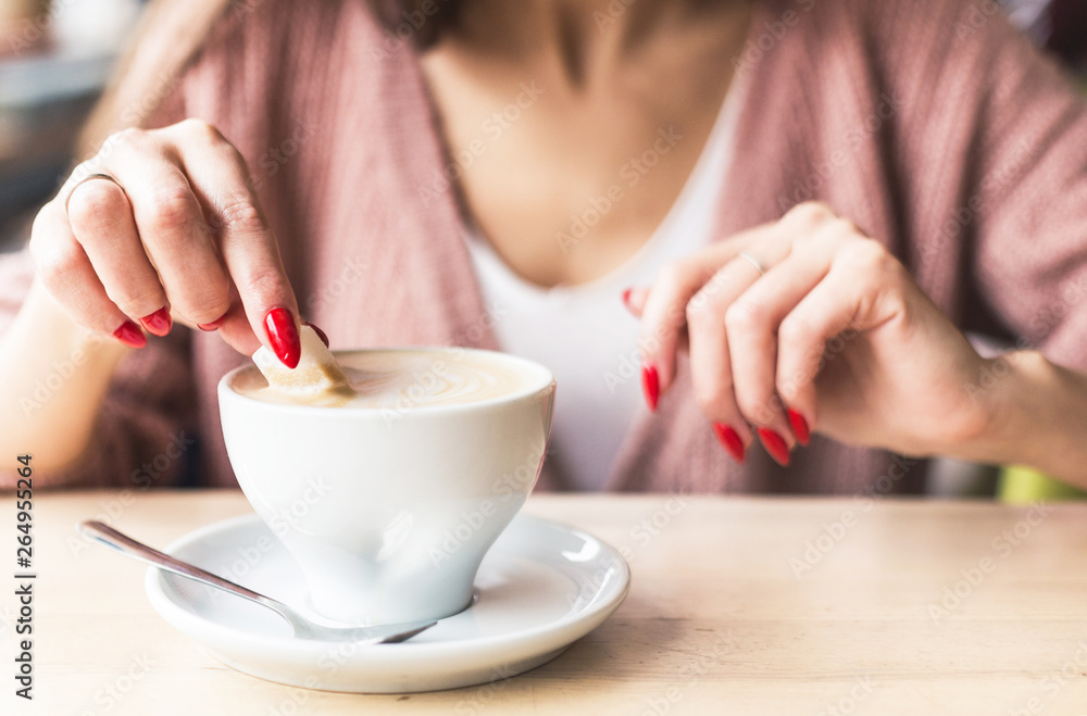 A girl dunks a piece of sugar in coffee. Closeup of hands with red nails make-up and cappuccino cup A girl dunks a piece of sugar in coffee. Closeup of hands with red nails make-up and cappuccino cup
