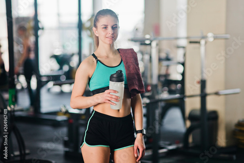 Fit woman with a towel on her shoulder and a bottle of water in her hand resting in the gym. Break time