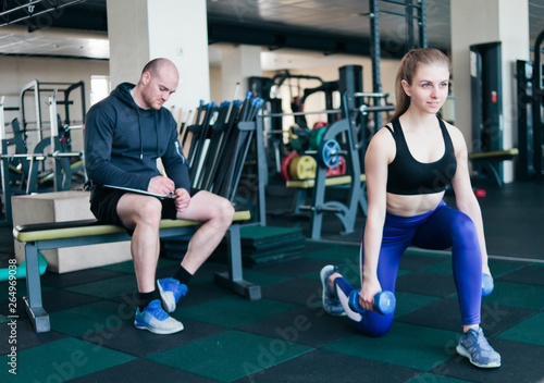 Fitness instructor supervises and notes in notebook results of training young athletic blonde performing exercises lunges with dumbbells in her hands in the gym