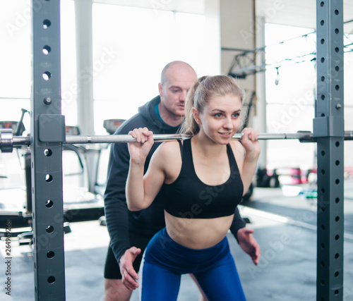 Young sportive woman crouches with a barbell on her shoulders with the help of a male instructor in the gym