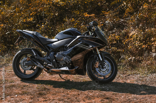 Dark Black sport motorcycle without logo and rider standing on the grass in forest on sunny day. Power motorbike parking on the meadow on sunset. Black super sport moto - photo from profile (side).