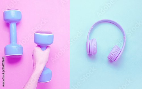 Creative concept fitness. Female hands hold plastic blue dumbbells, headphones on blue pink pastel background. Minimalism, Duotone effect