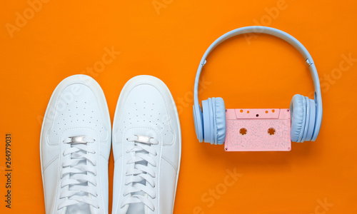Retro style. 80s. Pop culture. Minimalismalism. Headphones with audio cassette, white sneakers on orange background. Top view