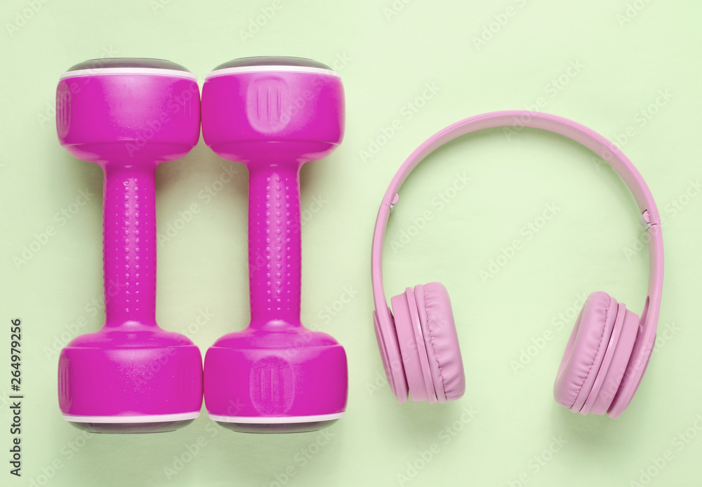 Pink headphones, plastic dumbbells on green pastel background. Sports with music. Top view.