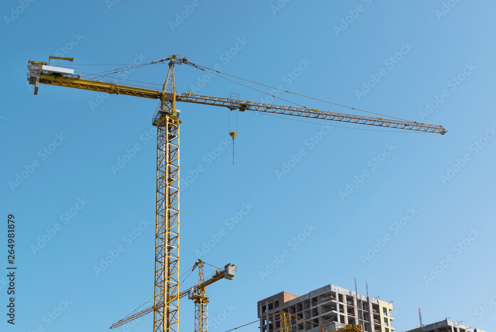 high-rise construction crane with a long arrow of yellow color against the blue sky over a new multi-storey building of concrete and brick under construction