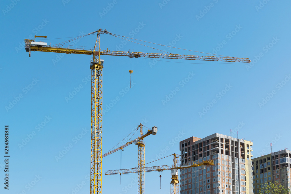 high-rise construction crane with a long arrow of yellow color against the blue sky over a new multi-storey building of concrete and brick under construction