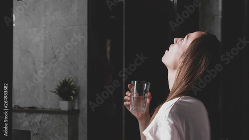 Young woman rinses her mouth with water while looking at mirror and spits water into the sink, side view.