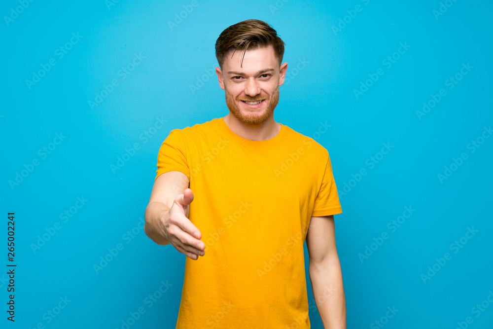 Redhead man over blue wall shaking hands for closing a good deal Redhead man over blue wall shaking hands for closing a good deal