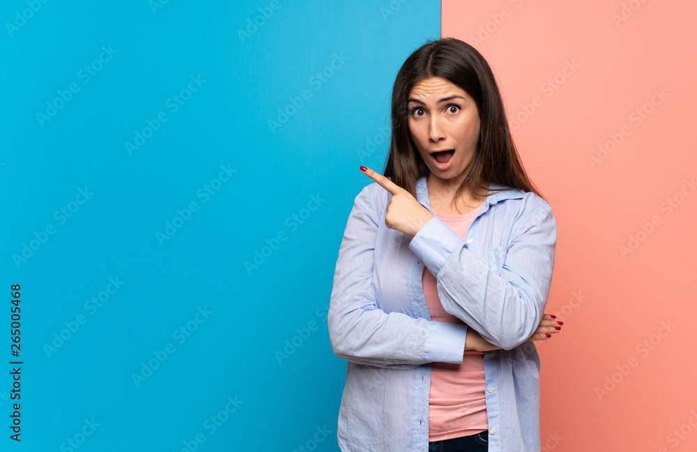 Young woman over pink and blue wall surprised and pointing side Young woman over pink and blue wall surprised and pointing side