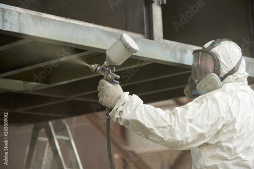 A man in a white uniform applies paint with a spray gun on a metal product.