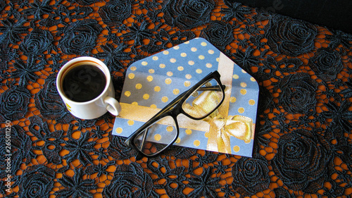 A cup of coffee, glasses and a greeting card on a tablecloth with black openwork ornament.