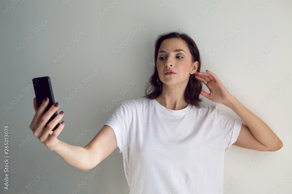 girl in a white t-shirt takes a selfie on a black phone against the white wall girl in a white t-shirt takes a selfie on a black phone against the white wall