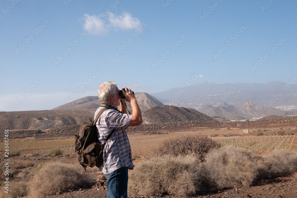 Side view of the senior man with backpack looking with a binoculars the landscape around. Volcanic island and arid climate. Healthy physical exercises