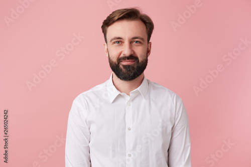 Portrait of young calm smiling handsome bearded man, wearing a white shirt. Looking at the camera isolated over pink background.