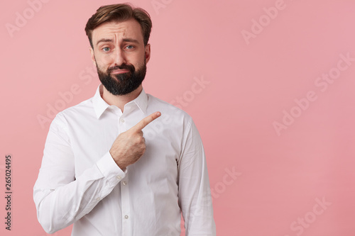 Frowning unhappy man wants to draw your attention pointing with finger on the copy space on the right, isolated over pink background.