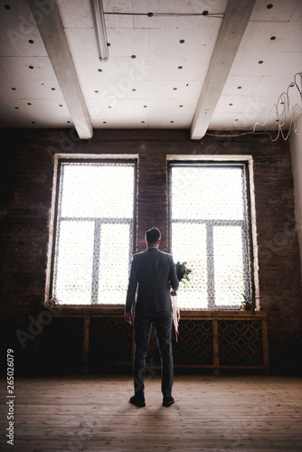 The groom is waiting for the bride in a gray suit. Groom holding bouquet and waiting for bride. Wedding day. The groom stands near a large window.