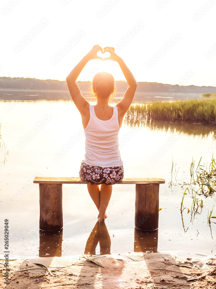 A young girl sits on a bench by the river enjoys a beautiful sunset and shows hands fingers heart sign. Girl sitting near water outdoors. Golden sunset on the lake. Young woman thinking about