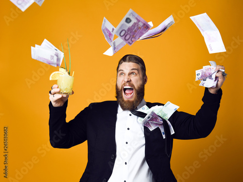 Happy young businessman with glass of cocktail in formal clothes holding bunch of money banknotes