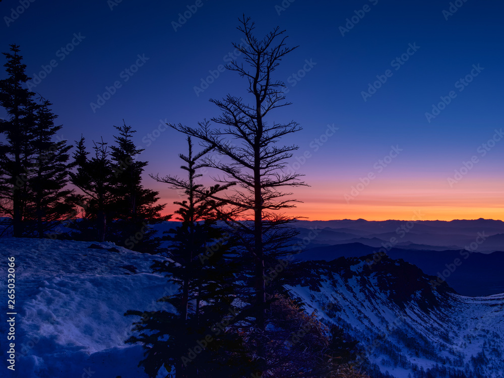 山頂にて朝焼け前の風景、雪景色で木々かシルエットになり、水平線の山々が朝焼色にピンク色に染る。