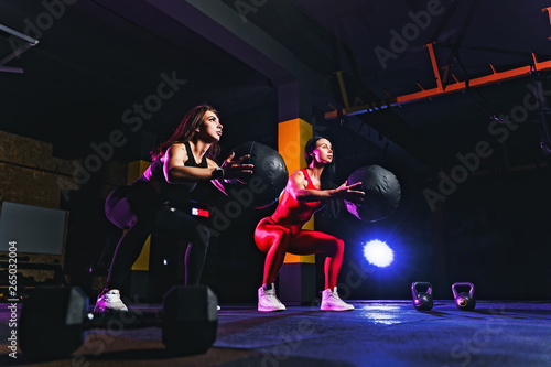 Two Sports women doing squat exercises with fitness ball. Female exercising and stretching with medicine ball at gym.