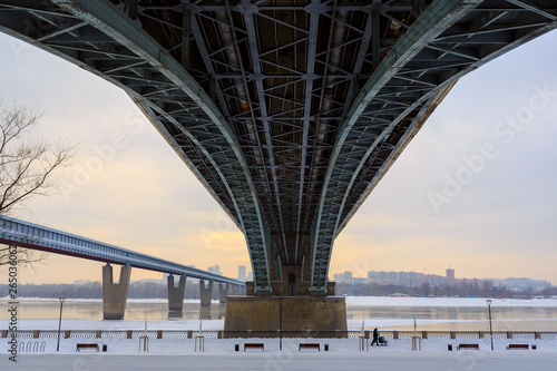 October bridge over the Ob River in Novosibirsk, Siberia, Russia. Bottom view on large metal structures. People walk along the snow-covered embankment. In the distance Metro Bridge and buildings.