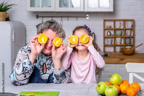 Happy little girl and her grandmother have breakfast together in a white kitchen. They are having fun and playing with fruits. Maternal care and love. Healthy eating