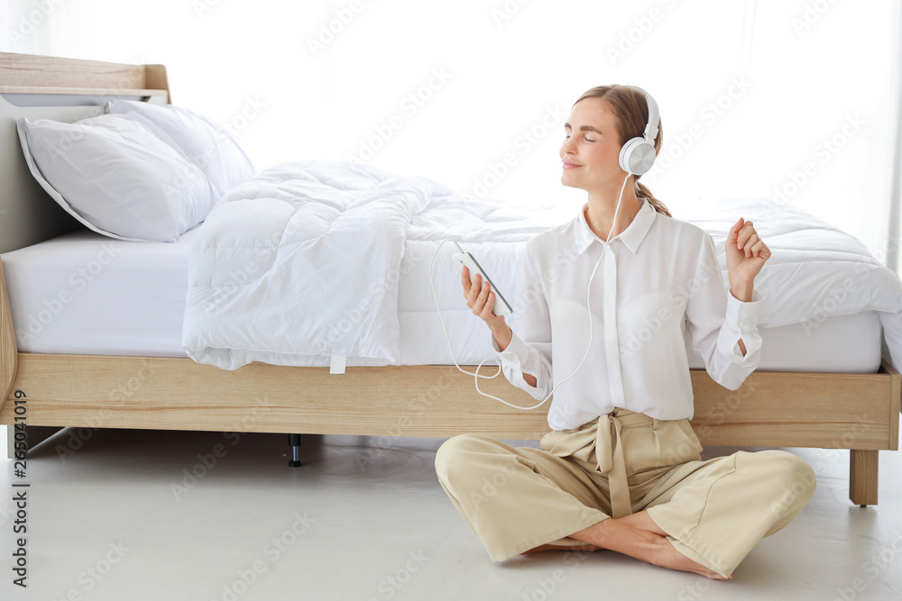 Young attractive caucasian woman wearing white casual clothes with headphone. She listening music or radio by using cell phone or music player in bedroom. (relaxation with technology concept) Young attractive caucasian woman wearing white casual clothes with headphone. She listening music or radio by using cell phone or music player in bedroom. (relaxation with technology concept)
