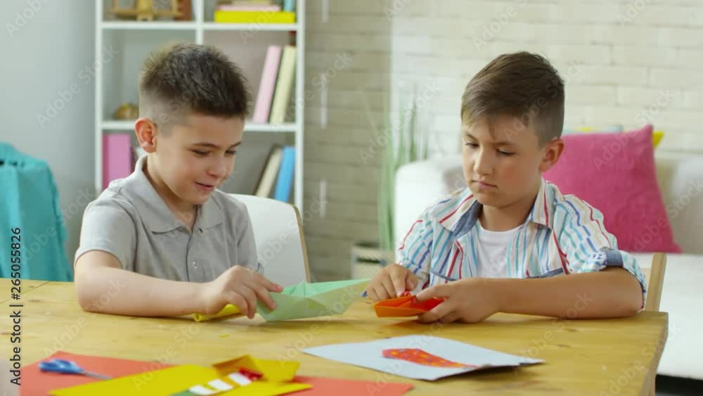 Two little caucasian boys sitting at desk in art class and discussing handmade origami boats Two little caucasian boys sitting at desk in art class and discussing handmade origami boats