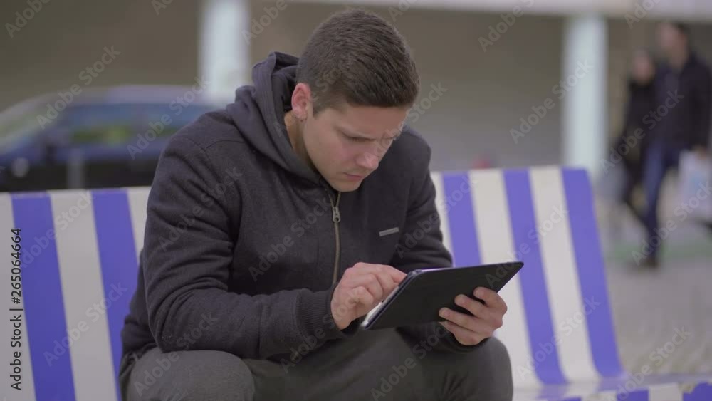 Serious man using digital tablet on street. Handheld shot of concentrated young man sitting on bench and using tablet pc outdoor. Wireless technology concept Serious man using digital tablet on street. Handheld shot of concentrated young man sitting on bench and using tablet pc outdoor. Wireless technology concept
