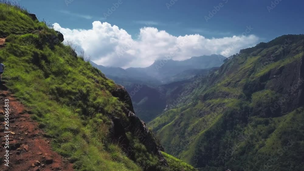 Woman walks on the mountain trail lying on the edge of the cliff with stunning valley views. The place named Little Adam's Peak in Ella, Sri Lanka Woman walks on the mountain trail lying on the edge of the cliff with stunning valley views. The place named Little Adam's Peak in Ella, Sri Lanka
