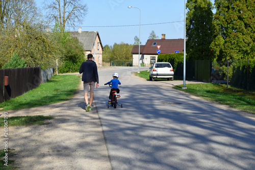 Boy in safety helmet cicling first bike on road in spring day  father learning little son to ride with bicycle on street  back view. Happy child making sports. Active leisure activities for children.
